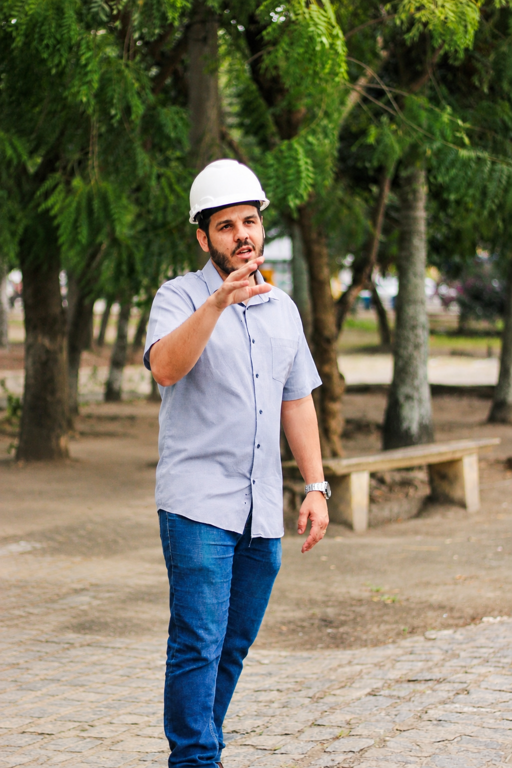 Joab Machado, de camisa azul e capacete branco, gesticula em um ambiente externo arborizado.